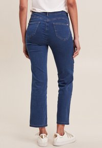 Jeans in denim blu scuro con taglio dritto, dotati di due tasche posteriori e cuciture a contrasto. Indossati con sneakers bianche.