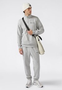 Felpa grigia con zip e pantaloni abbinati, entrambi con un logo. Indossato con un cappellino di lana bianco e una borsa a tracolla beige.