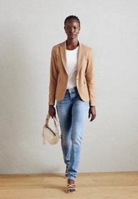 Béžový blazer, bílý žebrovaný top, světle modré džíny a béžové sandály na řemínky. Modelka drží malou, světle zbarvenou kabelku. Jednoduché pozadí.