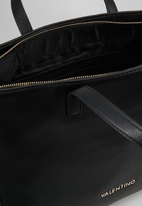 Svart läderväska med texturerad yta, guldräls, och satinfodrad insida. Har dubbla handtag och en logotypdetalj på framsidan.