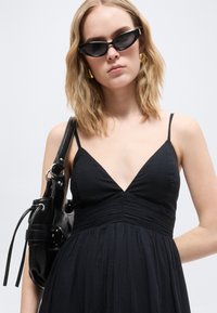 Γυναίκα με μαύρο αμάνικο φόρεμα, μαύρη τσάντα ώμου, γυαλιά ηλίου με μαύρο πλαίσιο σε σχήμα γατιού και χρυσούς κρίκους σκουλαρίκια μπροστά σε απλό φόντο.