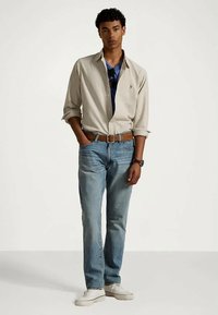 Lys beige langermet skjorte, blå grafisk T-skjorte under, lyseblå jeans, brunt belte, hvite joggesko, stående posisjon mot en ensfarget bakgrunn.