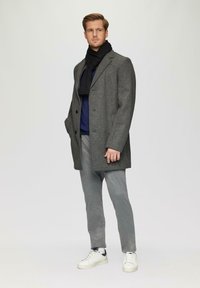 Grau karierter Mantel mit schmalem Revers, über einem Marineblauen Pullover getragen, kombiniert mit grauen Hosen und weißen Sneakers, akzentuiert durch einen schwarzen Schal.