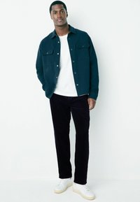 Dark green shirt jacket with two chest pockets, white t-shirt underneath, navy corduroy pants, and white sneakers with beige soles.