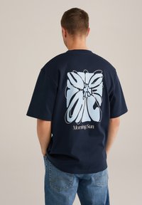 Sötétkék pamut póló rövid ujjakkal, amelyen egy nagy világoskék virágminta és a "Reggeli Nap" felirat található a hátsó oldalon.