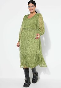 Groen patroon jurk met doorschijnende lange mouwen, getailleerde taille en gelaagde zoom. Gecombineerd met zwarte kniehoge laarzen met riemen.