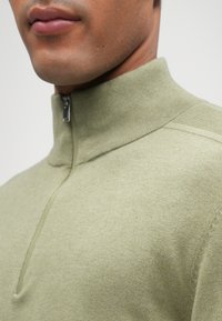 Grön zip-up tröja med hög krage, tillverkad av mjukt tyg. Har en strukturerad design och subtila ribbade detaljer vid kragen.