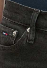 Fekete farmernadrág sima textúrával, ezüst gombbal, varrott derékpánttal és a zseb mellett egy kis háromszínű logóval.