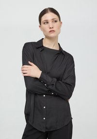 Camicia nera a maniche lunghe con apertura a bottoni, realizzata in tessuto liscio; presenta un colletto, orlo arrotondato e bottoni bianchi.