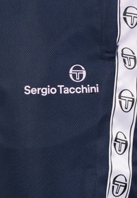 Maritieme sportkleding met een gestructureerde stof. Opvallend logo in het wit: "Sergio Tacchini." Zijstrepen met herhaald logo patroon.