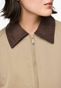 Bēšā jaka ar brūnu korduroja apkakli, kas ir aizsēta, rādot daļu no personas kakla un žokļa, kas novērsies sānis.