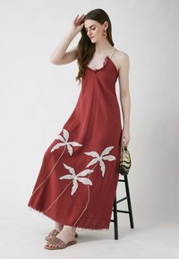 Rotes Maxikleid mit weißen, bestickten Palmblattmustern. Mit Spaghettiträgern und einem fransigen Saum. Tragend mit beigen Slippers.