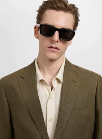 Blazer maroniu texturat peste o cămașă bej, purtat cu ochelari de soare negri. Accent pe croiala structurat și paleta de culori neutre.