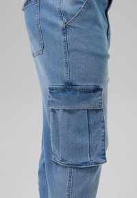 Hellblaue Denim-Cargo-Hosen mit einer Seitentasche, detaillierter Nahtführung und einem lässigen Schnitt. Der Stoff hat eine glatte Textur.