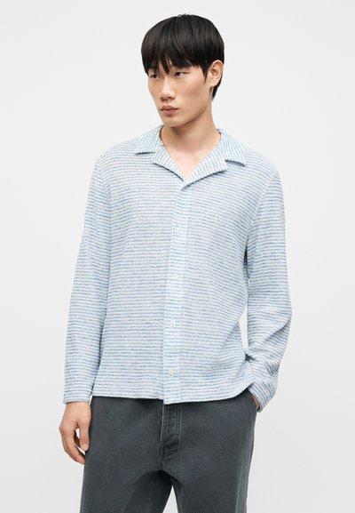 Chemise à rayures bleu clair et blanc avec col pointu, manches longues et coupe décontractée, associée à un pantalon gris.