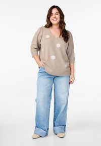 Maglione beige a maglia con ricami floreali, scollatura a V e maniche a palloncino corte. Abbinato a jeans beige a gamba larga e scarpe beige.