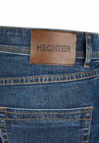 Denim jeans met een donkere wassing, met een bruine leren patch met het label "HECHTER" en contrasterende gele stiksels langs de tailleband en zakken.