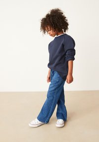 Marineblauwe sweatshirt met opgerolde mouwen, gecombineerd met losse blauwe jeans en witte sneakers. Achtergrond is neutraal en glad.
