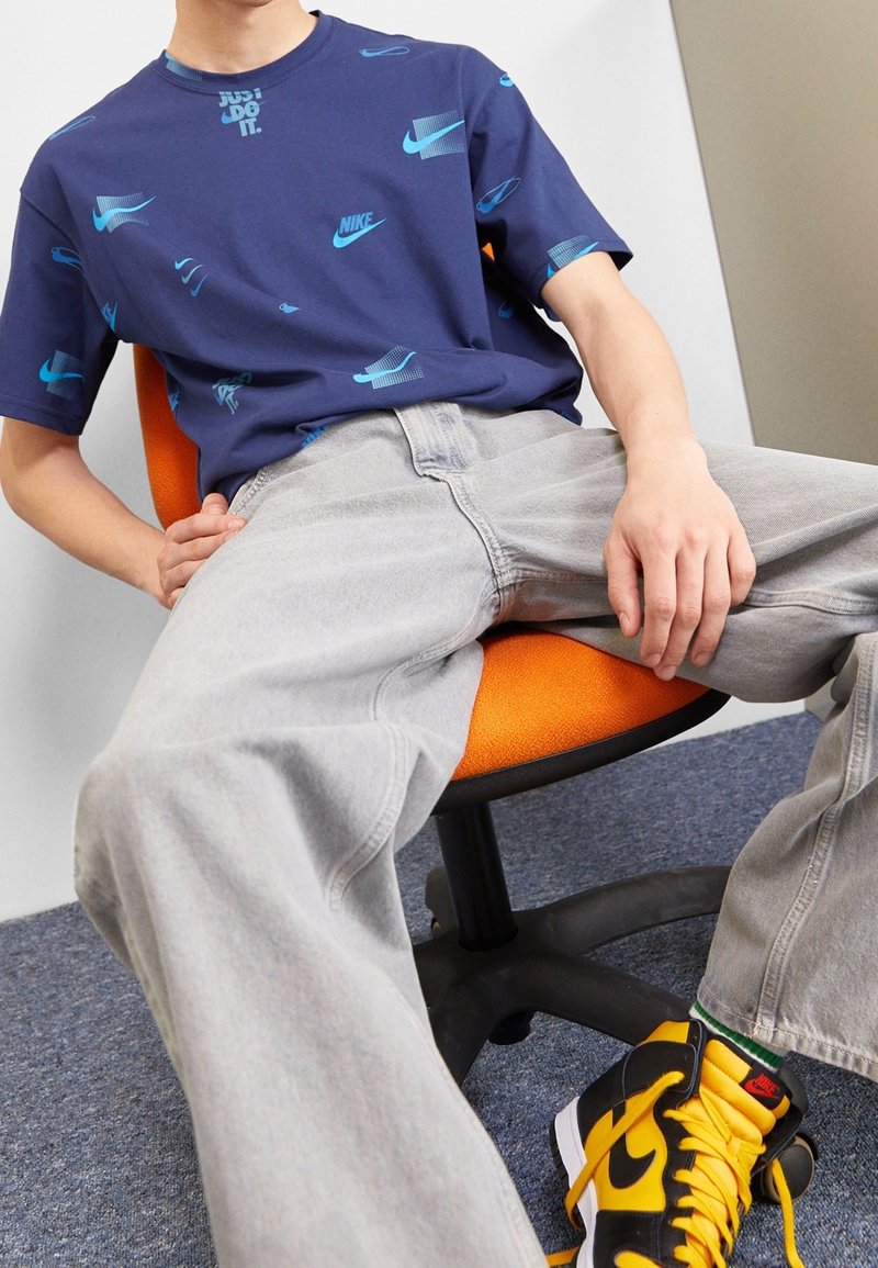 Tmavě modré bavlněné tričko s opakujícími se světle modrými logy Nike, spojené se světle šedými džíny a černo-žlutými teniskami Nike. Sedí na oranžové židli.