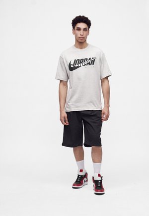 Νέος άνδρας στέκεται φορώντας ένα ελαφρύ T-shirt Jordan Air, μαύρο σορτς, λευκές κάλτσες και κόκκινα, μαύρα και λευκά αθλητικά παπούτσια σε λευκό φόντο.