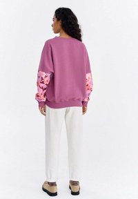 Roosa pusa lillemustriliste varrukatega, lõdva lõikega, soonikkoes mansettidega, kombineeritud kreemjate pükste ja massiivsete mustade jalanõudega.