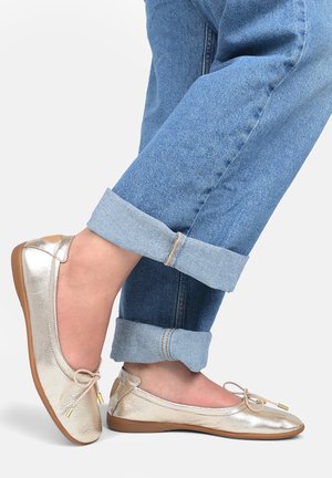 Ballerine metalliche oro con punta arrotondata, dettaglio a fiocco e suola in gomma tan flessibile; indossate con jeans in denim blu arrotolati.