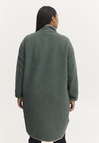 Abrigo de forro polar verde con cuello alto, hombros caídos y dobladillo redondeado. Presenta una textura esponjosa y un diseño hasta la rodilla.