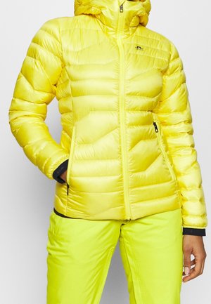 Kvinde iført en klar gul, quiltet hættejakke med lynlåslommer og matchende gule bukser mod en ensfarvet baggrund.