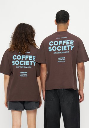 Dwie osoby stoją obok siebie, ubrane w brązowe koszulki z niebieskim napisem "Coffee Society, Slow Down, Brew Better, Caffeine Culture."