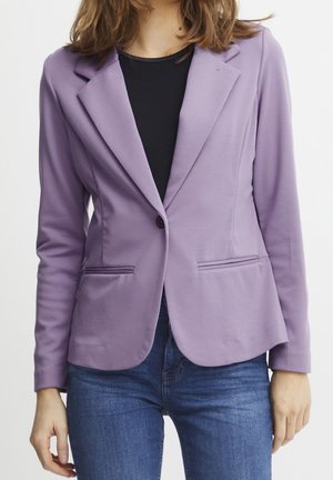 Mujer con una blazer ajustada color lavanda sobre una camiseta negra y vaqueros azules, de pie frente a un fondo blanco liso.