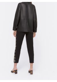 Vrouw in een zwarte leren jas, zwarte cropped broek met zijbloemversieringen en zwarte gestileerde muiltjes, staand tegen een witte achtergrond.