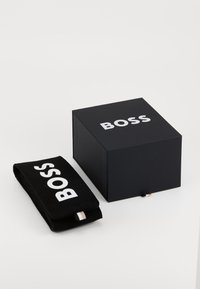 Boîte rectangulaire noire avec le logo "BOSS" blanc sur le dessus, accompagnée d'une pochette noire comportant le même logo. Les deux articles ont une texture lisse.