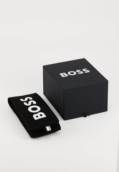 Черна правоъгълна кутия с бял логото "BOSS" отгоре, до черен джоб с едно и също лого. И двата предмета имат гладка текстура.