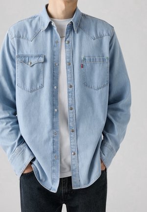 Homme portant une chemise en denim bleu clair boutonnée sur un T-shirt blanc, les mains dans les poches de son jean foncé, debout devant un fond uni.