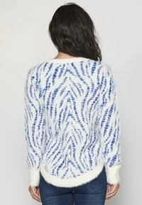 Fluffy white sweater featuring a blue abstract zigzag pattern, rounded hem, and long sleeves with ribbed cuffs, made from soft fabric.