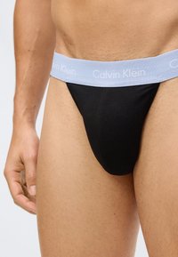 Svart bomullsstringtruse med lys blå elastisk midjebånd som har "Calvin Klein"-logo. Glatt tekstur, tettsittende passform, minimalistisk design.