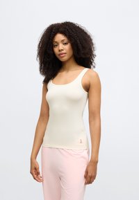 Šviesiai smėlio spalvos liemenėlė su apvalia iškirpte, pagaminta iš lygaus audinio, su mažu raudonu logotipu ant apačios, derinama su rožinėmis sportinėmis kelnėmis.