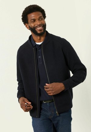 Smiling man wearing a dark zip-up jacket over a navy polo shirt and blue jeans, adjusting his jacket with both hands.