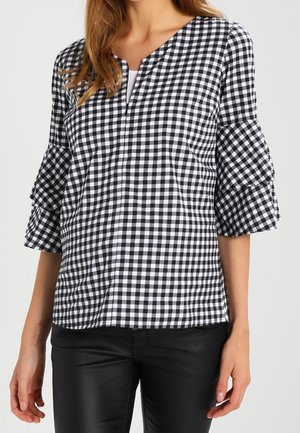 Kobieta w czarno-białej bluzce w kratkę vichy z falbanami na rękawach do połowy przedramienia oraz w czarnych dopasowanych spodniach.