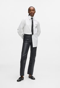 Λευκό πουκάμισο με μακριά μανίκια και μαύρη γραβάτα, συνδυασμένο με γυαλιστερά μαύρα παντελόνια και μαύρες μοκέτες. Το σύνολο διαθέτει μοντέρνα γραμμή και αντίθεση υφής.