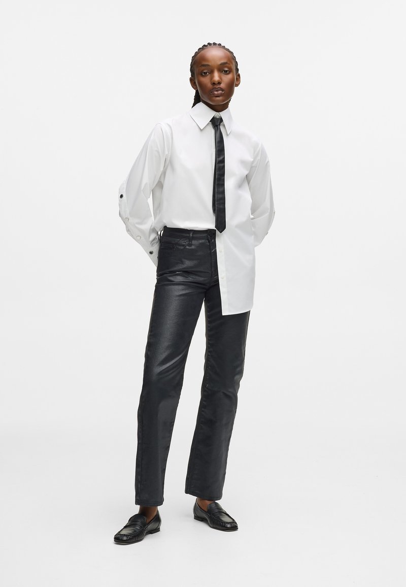 Λευκό πουκάμισο με μακριά μανίκια και μαύρη γραβάτα, συνδυασμένο με γυαλιστερά μαύρα παντελόνια και μαύρες μοκέτες. Το σύνολο διαθέτει μοντέρνα γραμμή και αντίθεση υφής.