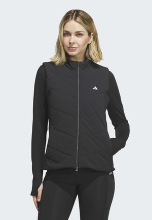 Frau mit schulterlangen blonden Haaren, die eine schwarze gesteppte Weste mit Reißverschluss über einem schwarzen Langarmshirt trägt und vor einem hellen Hintergrund steht.