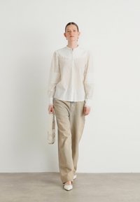 Camicia bianca con dettagli in pizzo sulle maniche, abbinata a pantaloni beige a gamba larga e scarpe bianche a punta, con una borsa piccola.