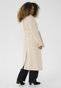 Kvinde med krøllet hår iført en beige bæltejakke, mørke vide bukser og sorte stiletter, der poserer mod en ensfarvet baggrund.