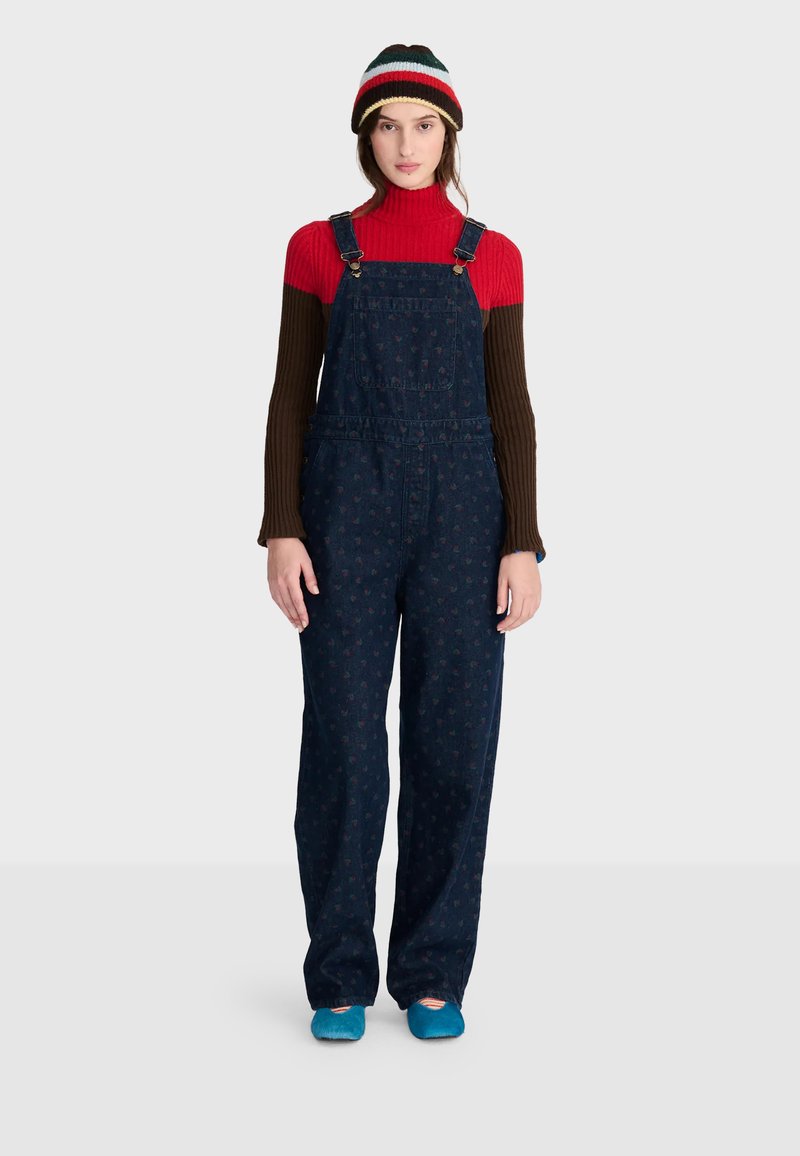Femme portant un bonnet tricoté multicolore, un pull à col roulé côtelé rouge et marron, une salopette bleue foncée à motifs et des chaussures bleues, debout sur fond blanc.