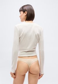 Γυναίκα με κοντά σκούρα μαλλιά, ντυμένη με μακρυμάνικη μπεζ μπλούζα και ταιριαστό στρινγκ, στραμμένη με την πλάτη προς ένα απλό ανοιχτό φόντο.