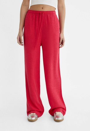 Femme portant un pantalon ample en lin rouge avec une taille élastique et un cordon de serrage, associé à des baskets argentées et un top court blanc.