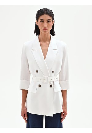 Donna con capelli neri corti effetto bagnato, indossa un blazer bianco doppiopetto con cintura e pantaloni scuri, in piedi su uno sfondo chiaro semplice.