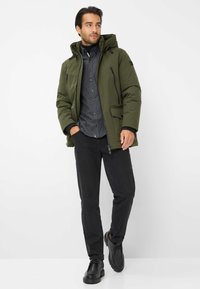 Giacca a soffietto verde oliva con cappuccio, cerniere nere e tasche laterali, indossata sopra una camicia grigia con bottoni e pantaloni neri, con scarpe nere.