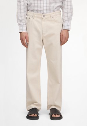 Mand iført beige bukser med lige ben, en ternet langærmet skjorte stoppet ind og sorte sandaler med åbne tæer, der står mod en hvid baggrund.